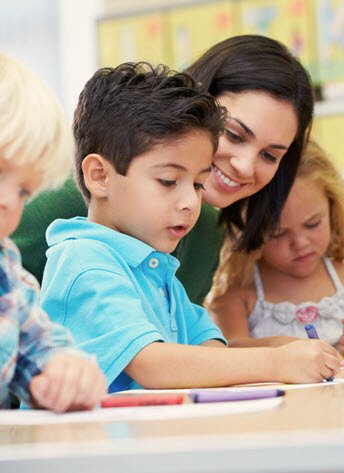 A smiling caregiver assisting a young boy in a blue shirt with his work at a classroom table, while other children are engaged in activities nearby.